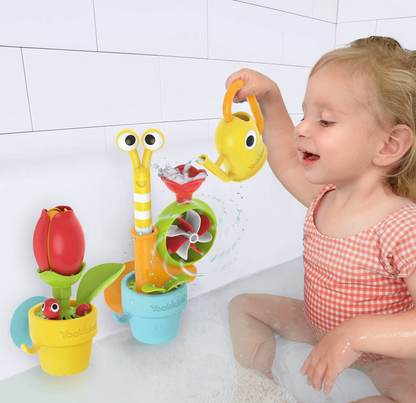 A toddler girl in a pink gingham swimsuit sits in a white tiled bathtub, playing with a colorful children's water toy set. The set includes a yellow flower-shaped watering can, a striped watering can, a green leaf spinner, and a red flower pot.
