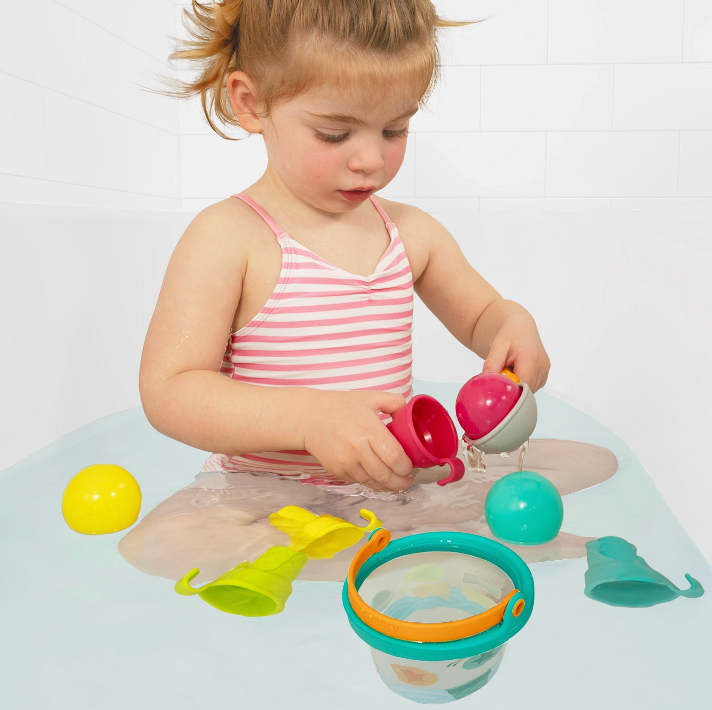 A young child sitting in a bathtub playing with a colorful bath toy set, including a pink and white striped tank top, a pink cup, a teal ball, and other bath accessories.