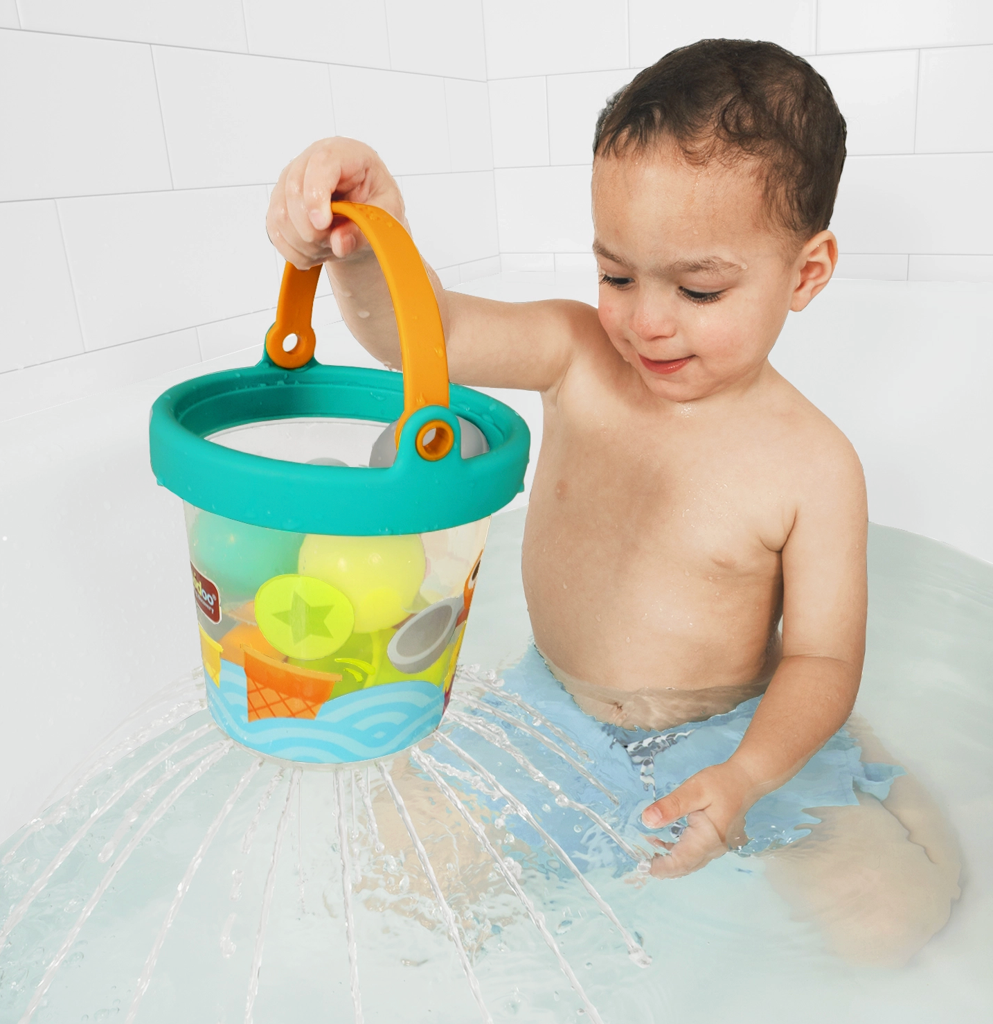 A toddler sitting in shallow water holds a colorful plastic bucket toy with an orange handle and turquoise rim. The bucket contains small beach-themed toys, and water sprays around the bucket against a white tiled wall.