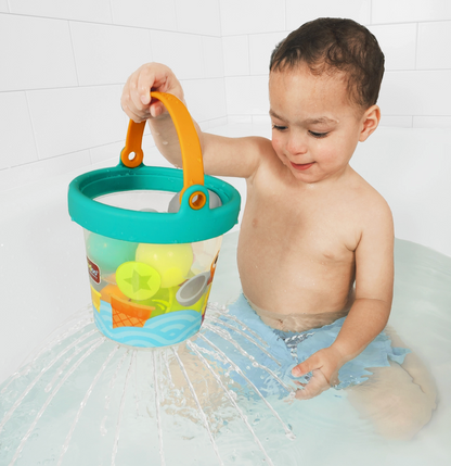 A toddler sitting in shallow water holds a colorful plastic bucket toy with an orange handle and turquoise rim. The bucket contains small beach-themed toys, and water sprays around the bucket against a white tiled wall.
