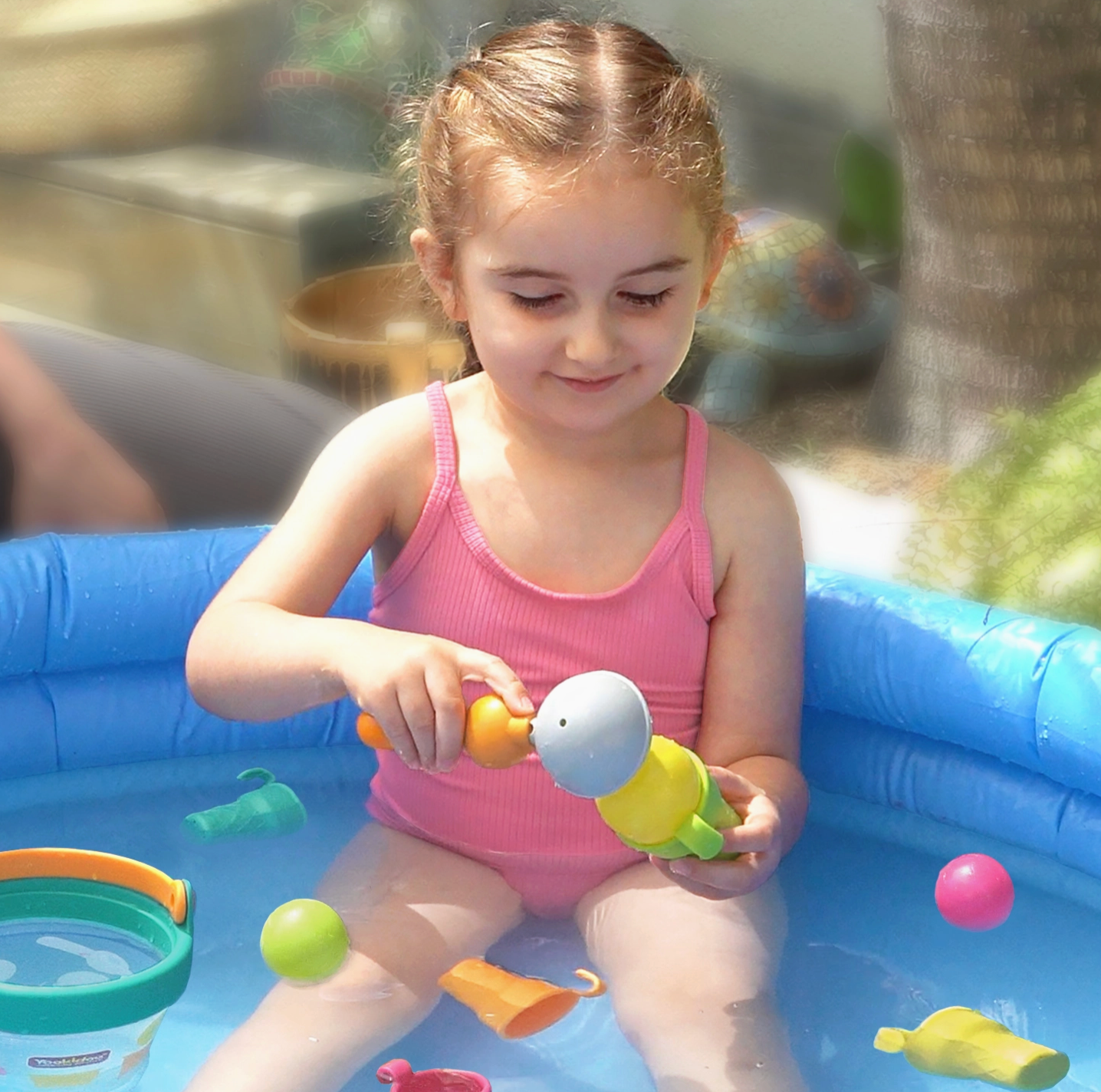 A young girl in a pink swimsuit sits in a blue inflatable pool, playing with colorful toy water fountains and balls outdoors.