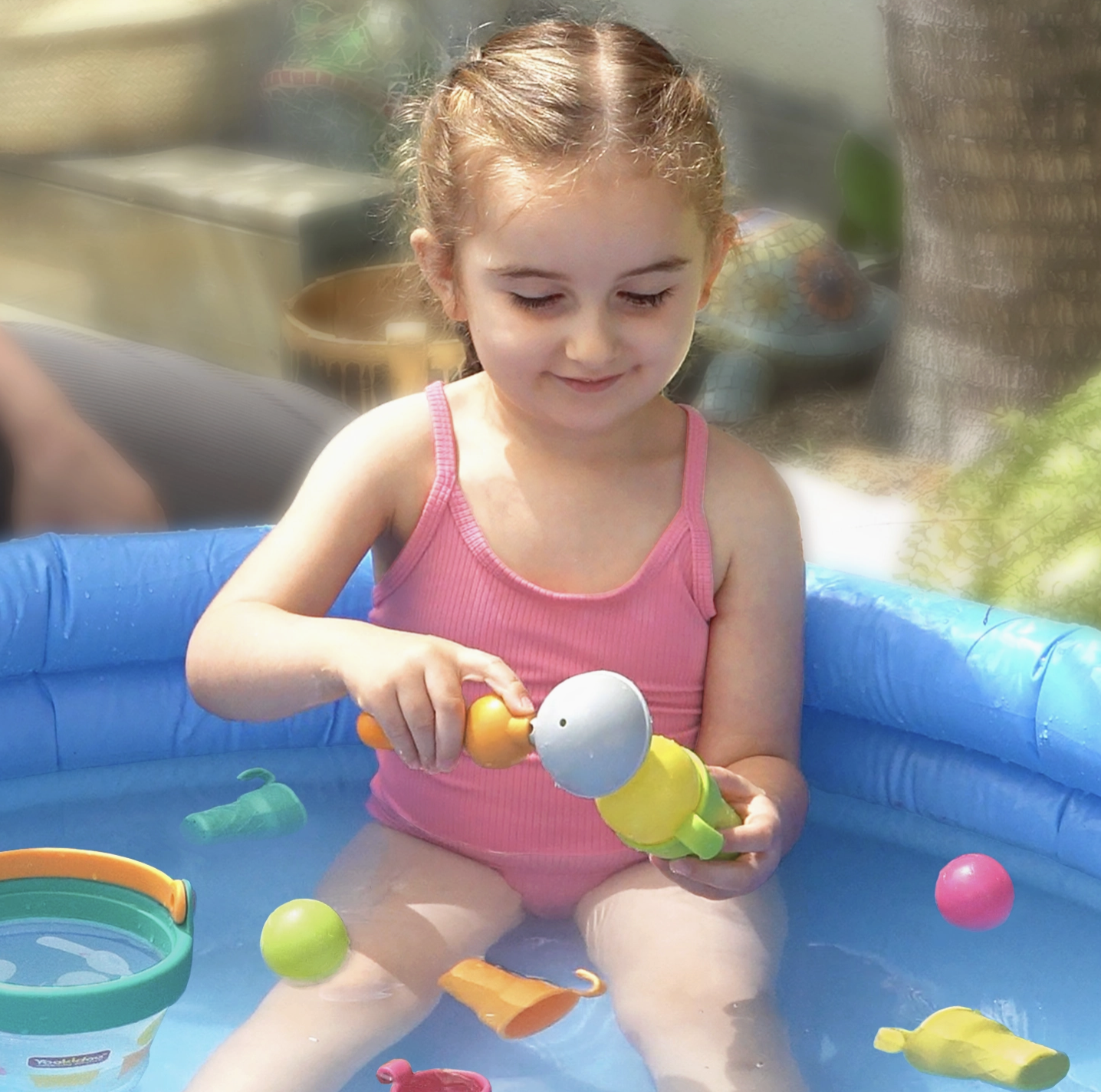 A young girl in a pink swimsuit sits in a blue inflatable pool, playing with colorful toy water fountains and balls outdoors.