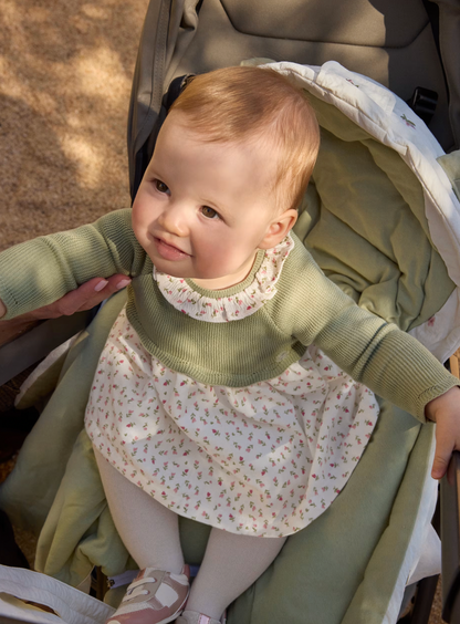 A baby wearing a green ribbed long-sleeve top and a cream floral dress sits in a stroller with a green blanket.