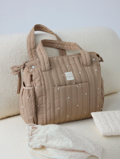 A quilted beige diaper bag with white polka dots sits on a cream textured sofa, with matching baby accessories placed in front.
