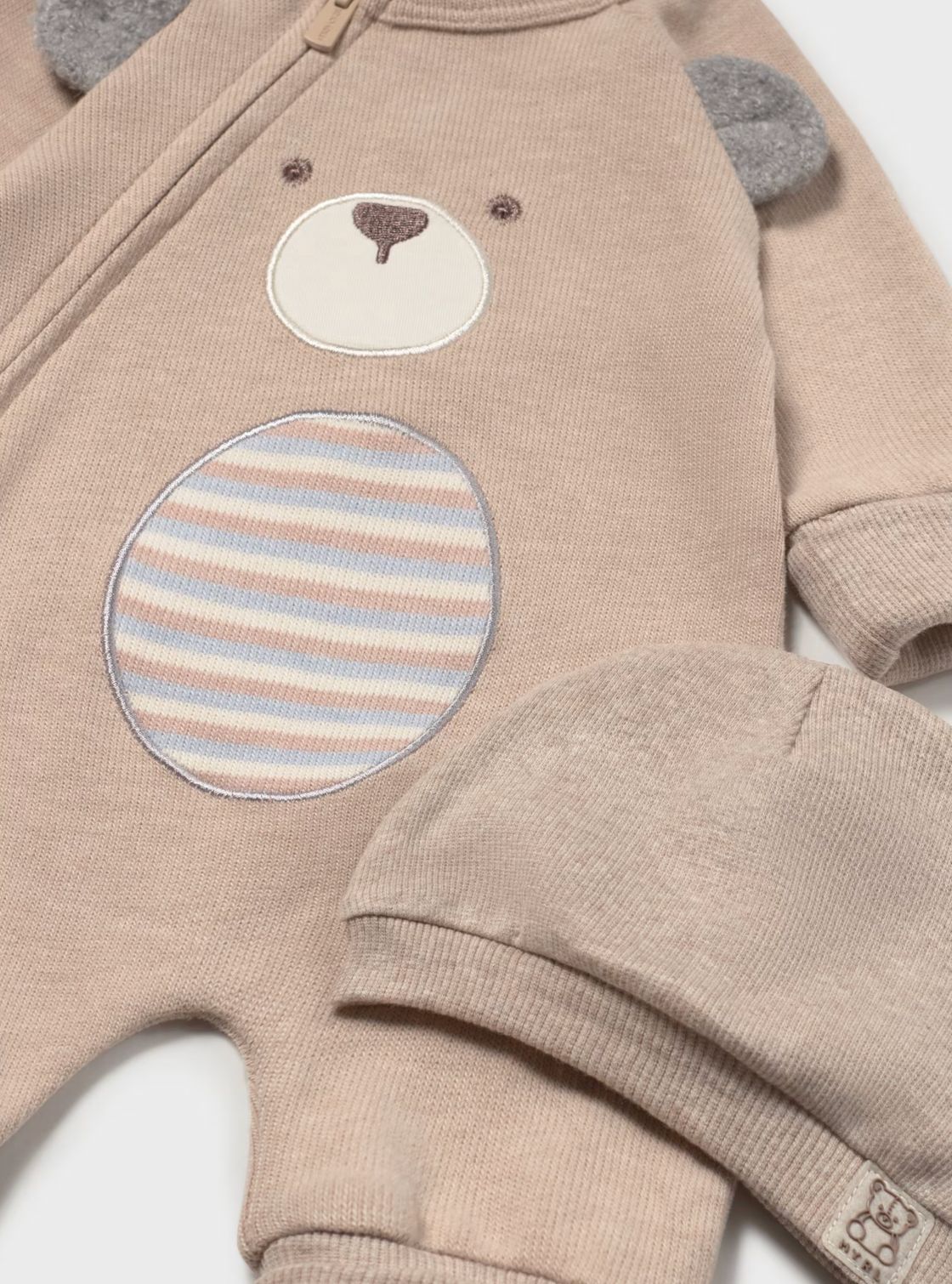 A close-up of a baby's long-sleeve knit romper in a warm beige/tan color, featuring an applique bear face on the chest with a striped circular patch and gray bear ears. A matching knit hat is partially visible in the foreground.