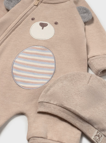 A close-up of a baby's long-sleeve knit romper in a warm beige/tan color, featuring an applique bear face on the chest with a striped circular patch and gray bear ears. A matching knit hat is partially visible in the foreground.