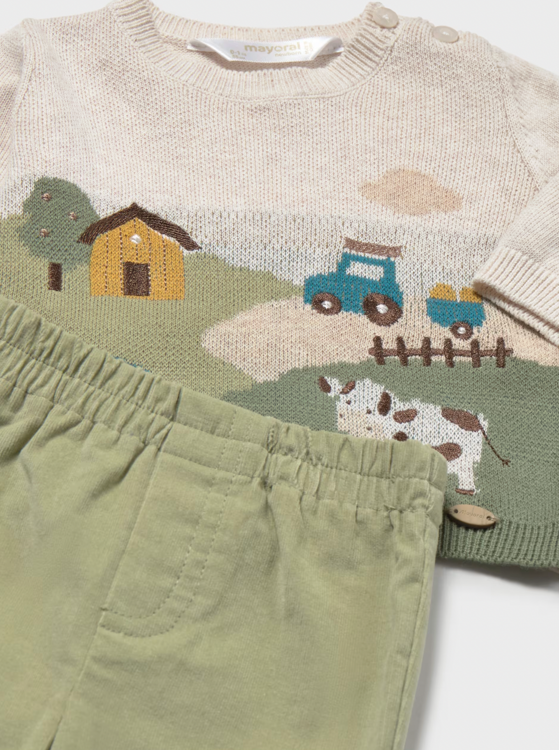 A baby knit sweater and matching pants set. The cream-colored knit sweater features a colorful hand-knit farm scene with a yellow house, green hills, a blue tractor, and a cow.