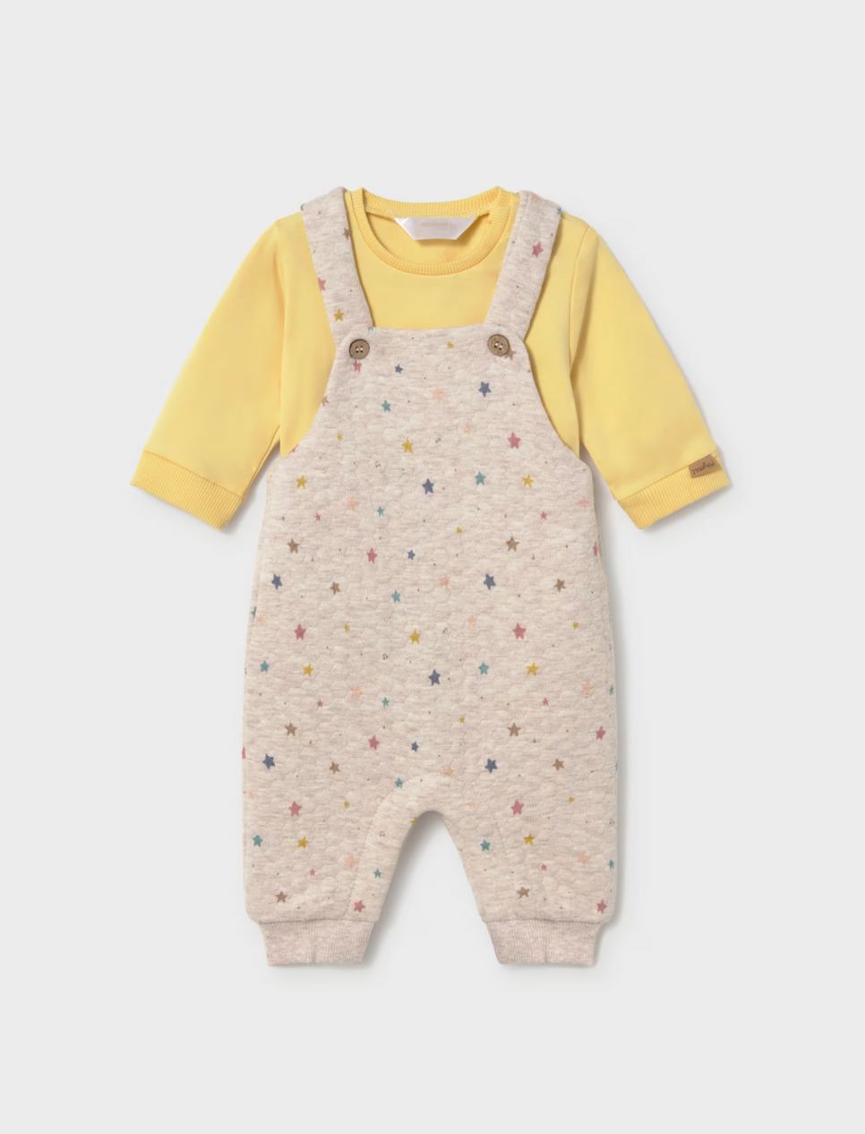 A baby's long-sleeve romper featuring cream-colored microfiber overall with a colorful star pattern, worn over a soft yellow long-sleeve shirt. The romper has adjustable shoulder straps with two brown buttons and ribbed cuffs and ankles.