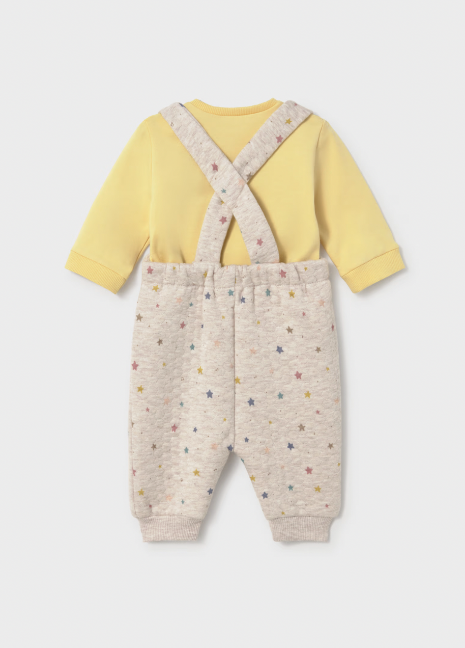 A baby two-piece outfit featuring a pale yellow long-sleeve top and light beige heathered pants with a multicolor star print and crossover suspenders, shown from the back on a light gray background.