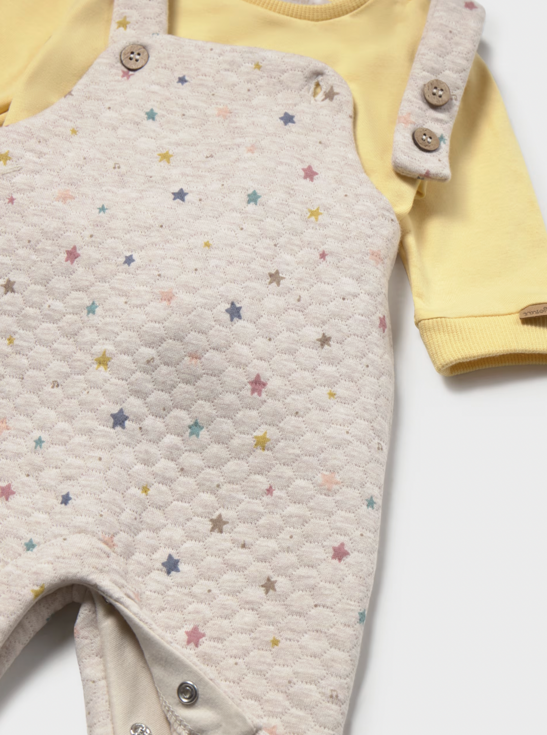 A baby's long-sleeve romper set featuring a cream-colored honeycomb quilted bib overlayer with a scattered pastel star pattern, worn over a soft yellow long-sleeve bodysuit. The bib has buttoned shoulder straps and snap closures at the crotch.