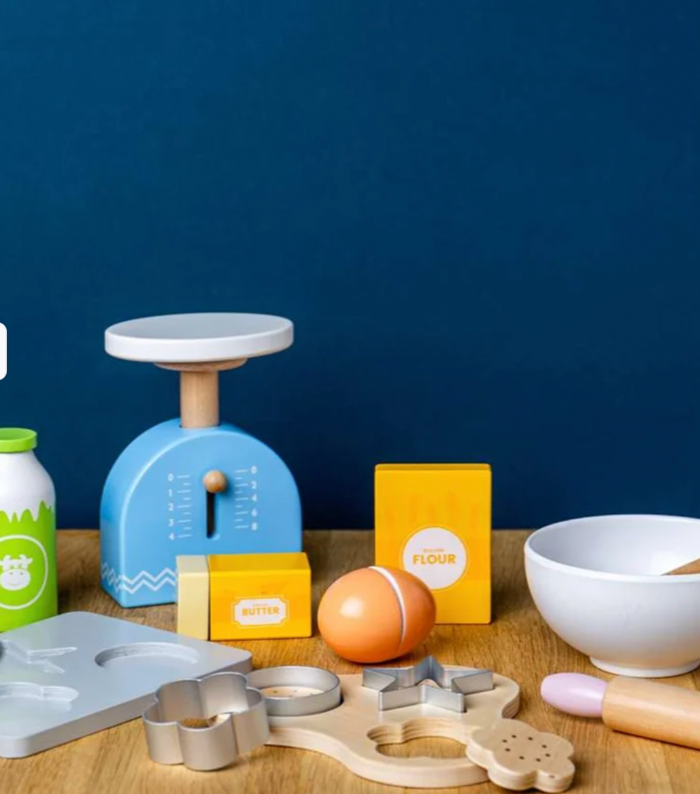 A children's wooden play kitchen set arranged on a tabletop against a dark blue background. The set includes a blue scale, yellow flour and butter boxes, an egg, a white bowl, a rolling pin, cookie cutters, and other wooden baking tools.