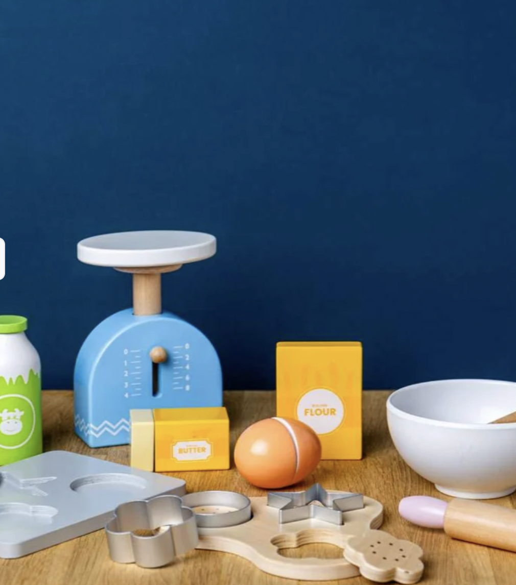 A children's wooden play kitchen set arranged on a tabletop against a dark blue background. The set includes a blue scale, yellow flour and butter boxes, an egg, a white bowl, a rolling pin, cookie cutters, and other wooden baking tools.