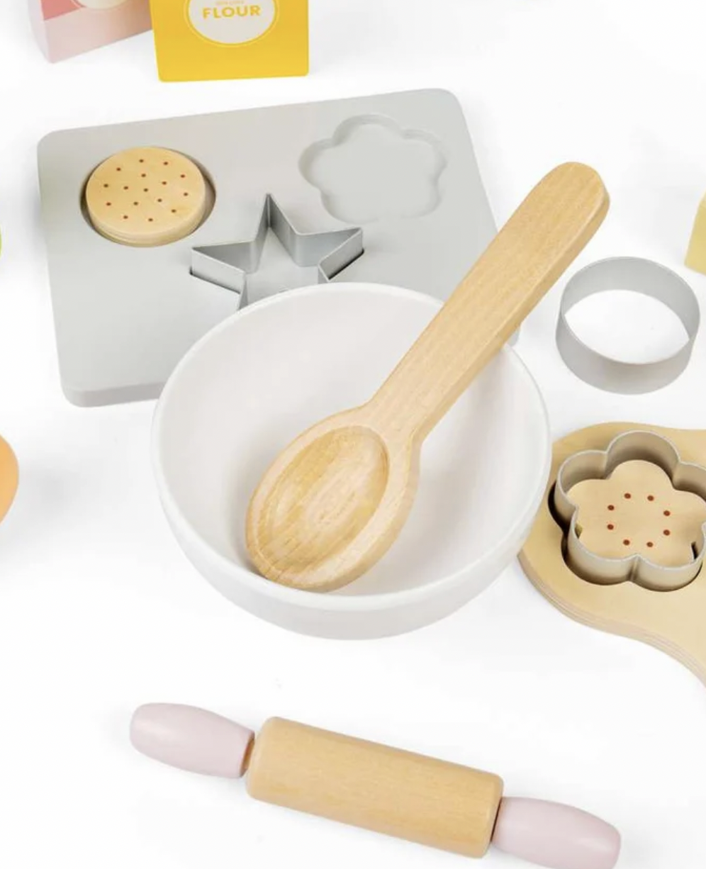 A children's wooden baking toy set arranged on a white background, including a light wood spoon in a white bowl, a wooden rolling pin, cookie cutters, a flour mold, and a pink-and-wood rolling cylinder.