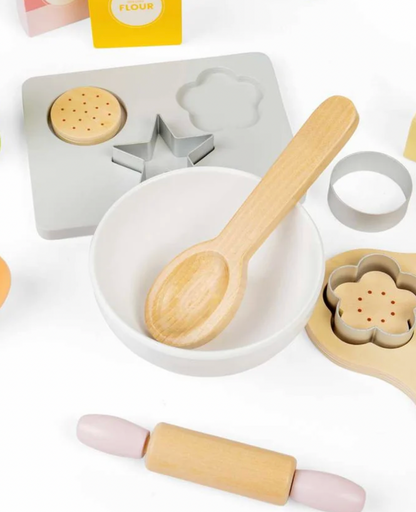 A children's wooden baking toy set arranged on a white background, including a light wood spoon in a white bowl, a wooden rolling pin, cookie cutters, a flour mold, and a pink-and-wood rolling cylinder.