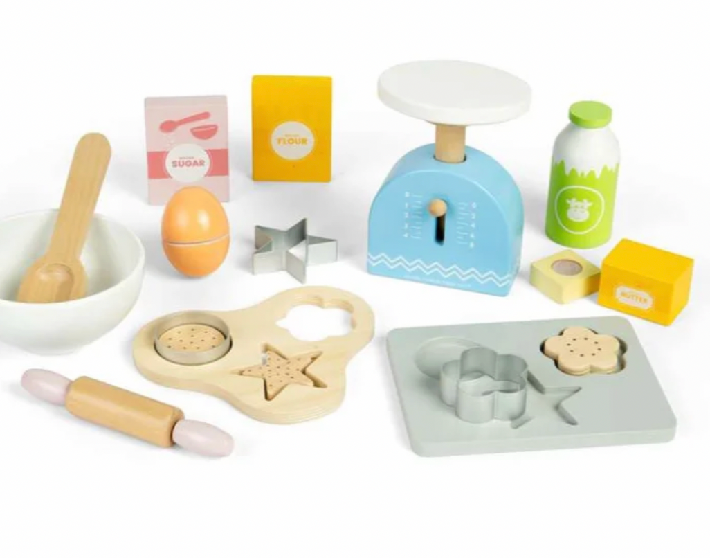 A children's wooden play kitchen set arranged on a white background, including a blue scale, milk bottle, sugar and flour boxes, bowl with spoon, rolling pin, cookie cutters, mold tray, egg, and other baking-themed pieces.