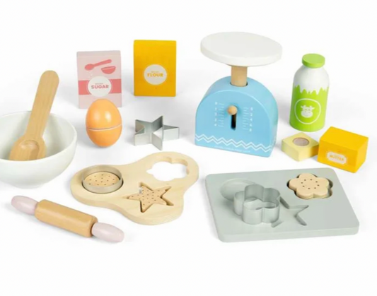 A children's wooden play kitchen set arranged on a white background, including a blue scale, milk bottle, sugar and flour boxes, bowl with spoon, rolling pin, cookie cutters, mold tray, egg, and other baking-themed pieces.