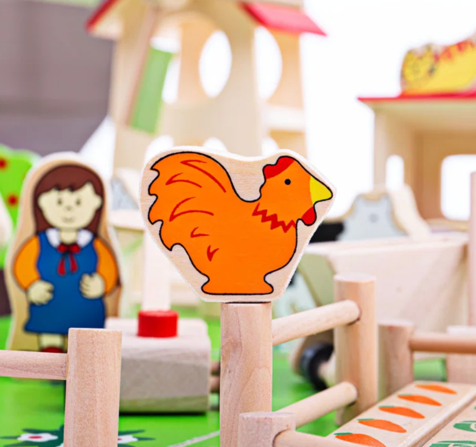 A wooden toy farm scene with a colorful orange rooster figure standing on a light wood fence, alongside other wooden toy pieces and a blurred playset background.