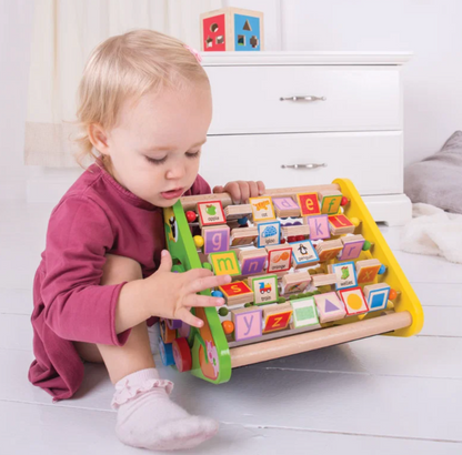 A toddler in a magenta long-sleeve shirt and light pink socks sits on a white wooden floor, interacting with a colorful wooden alphabet and number learning toy. The toy features a yellow side frame, green front edge, and rows of colorful alphabet blocks on a wooden base.