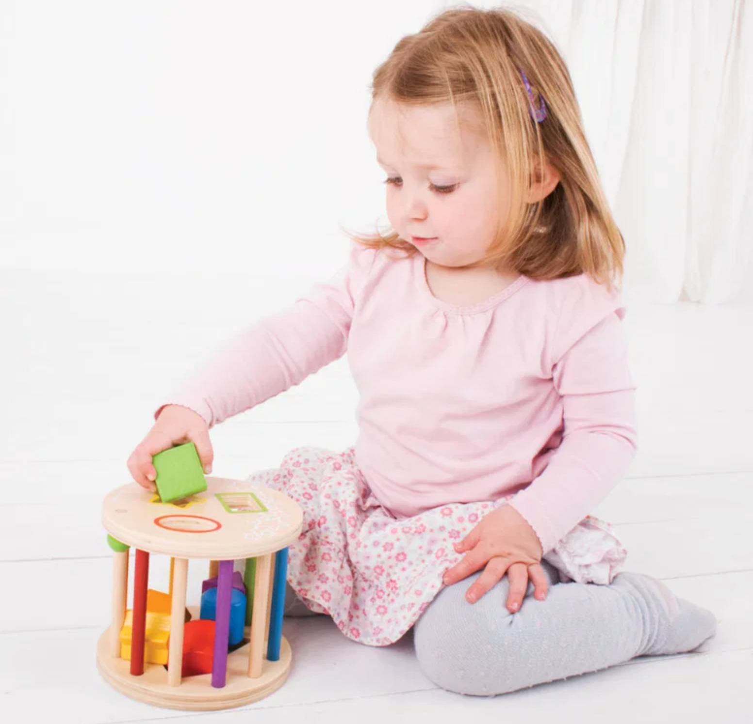 A young toddler girl with blonde hair sits on a white wooden floor, playing with a colorful wooden sorting toy. She is wearing a pink long-sleeve top, a pink and white floral skirt, and gray leggings.