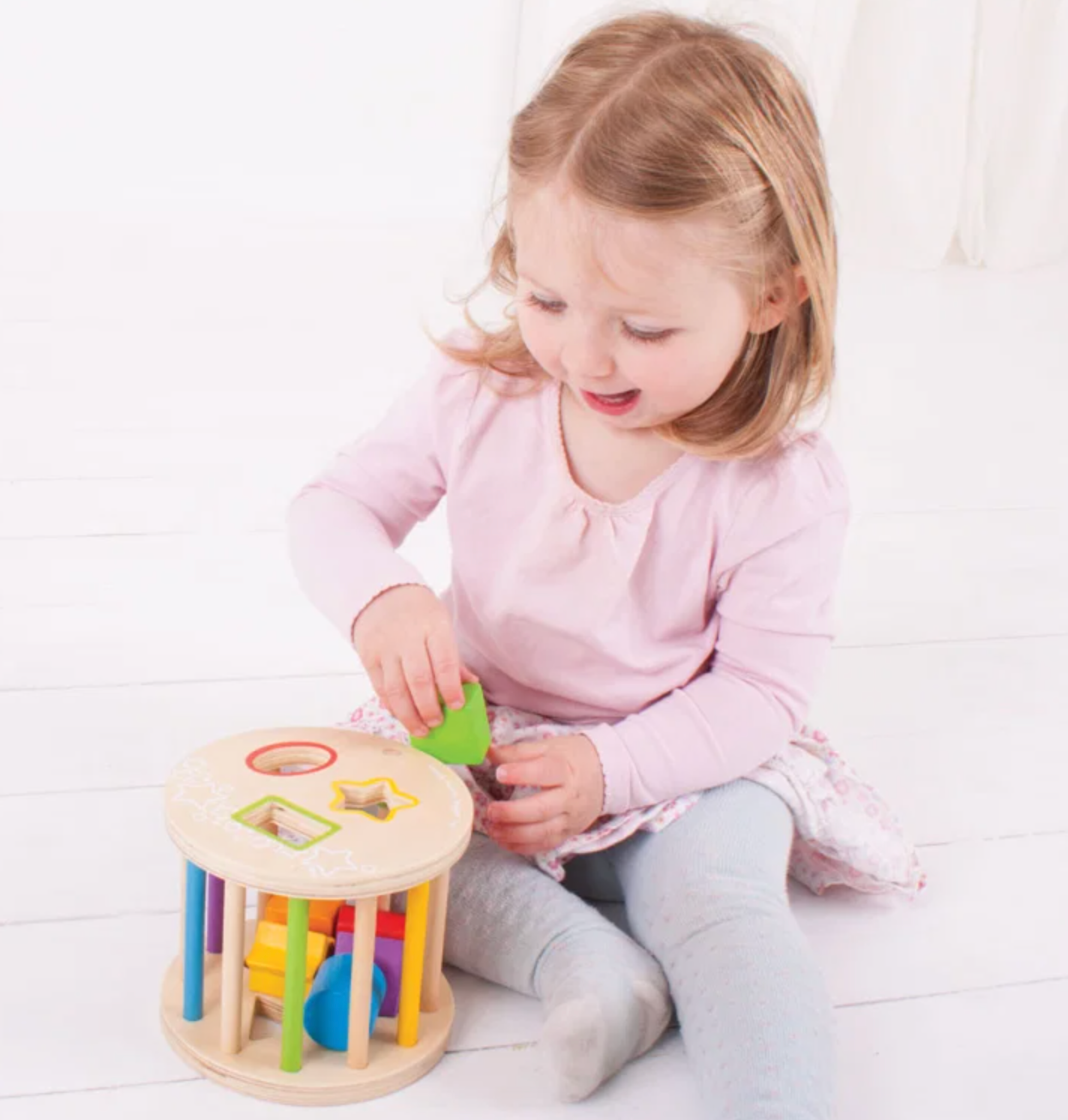 A young toddler girl with blonde hair sits on a white wooden floor, playing with a colorful wooden shape-sorting toy. She is wearing a light pink long-sleeve top and gray leggings.