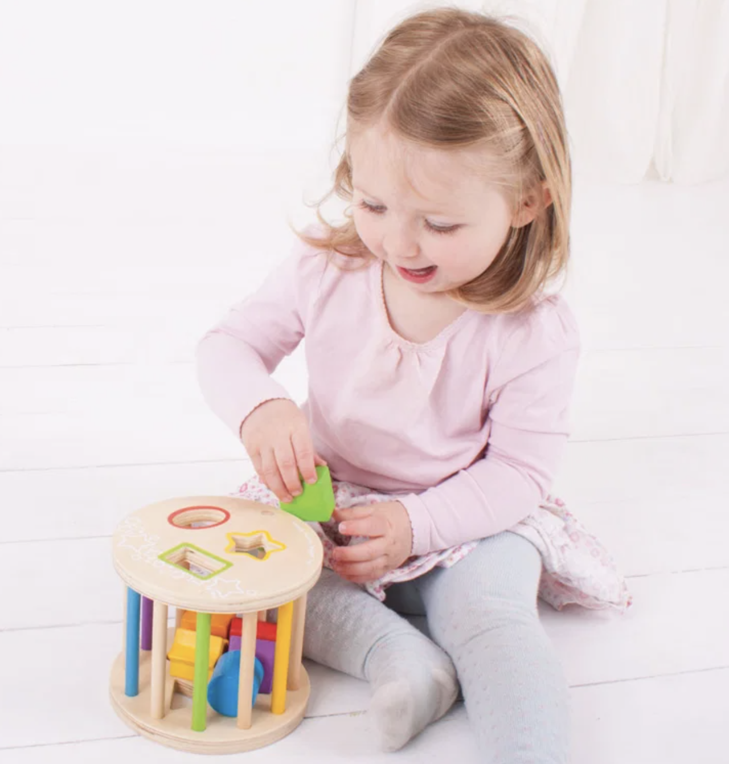 A young toddler girl with blonde hair sits on a white wooden floor, playing with a colorful wooden shape-sorting toy. She is wearing a light pink long-sleeve top and gray leggings.