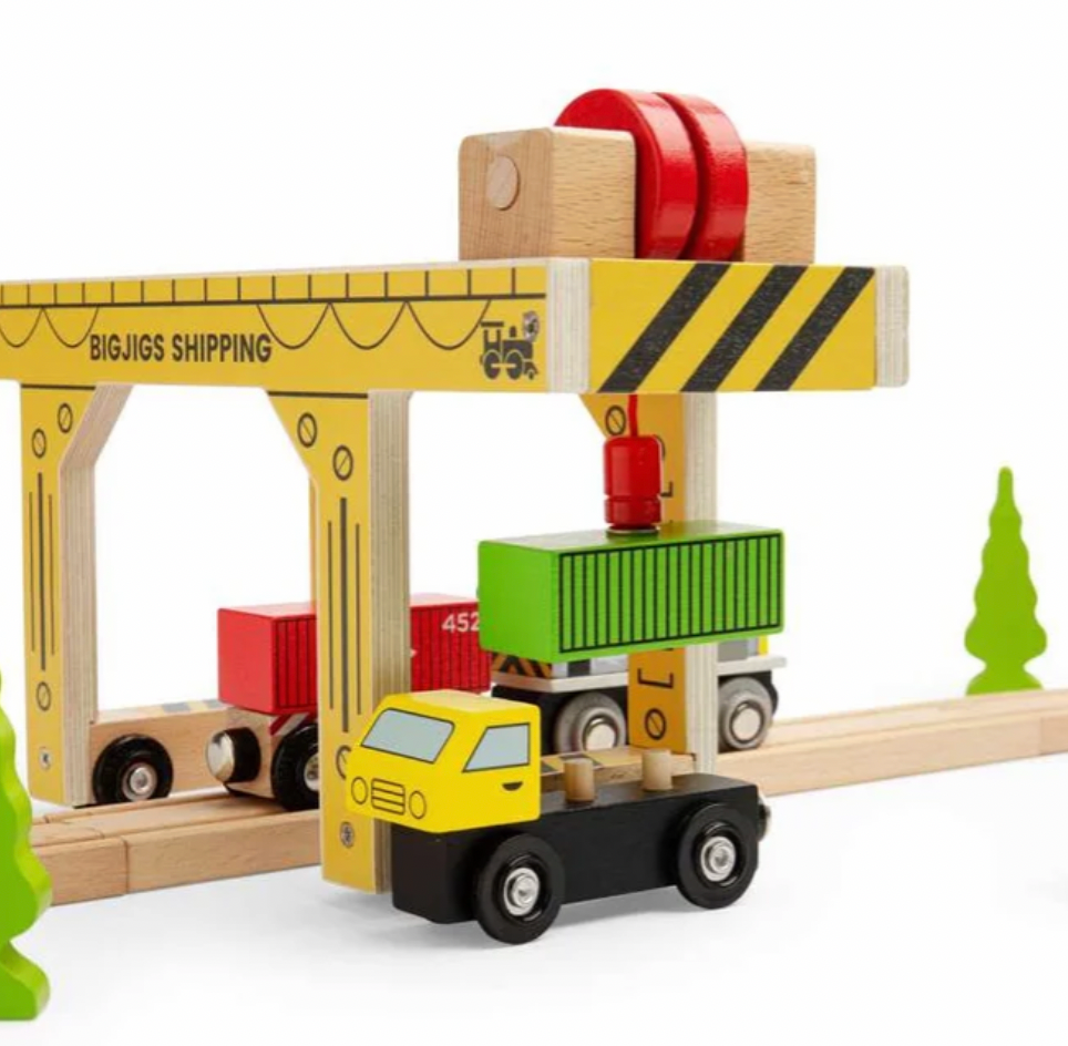 A wooden toy shipping crane and cargo truck set on a light wood railway track against a white background. The set includes a yellow crane labeled 'BIGJIGS SHIPPINGING', a yellow and black cargo truck carrying a green shipping container, and red and green cargo boxes.