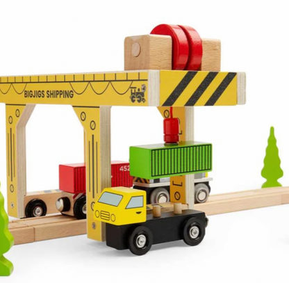 A wooden toy shipping crane and cargo truck set on a light wood railway track against a white background. The set includes a yellow crane labeled 'BIGJIGS SHIPPINGING', a yellow and black cargo truck carrying a green shipping container, and red and green cargo boxes.