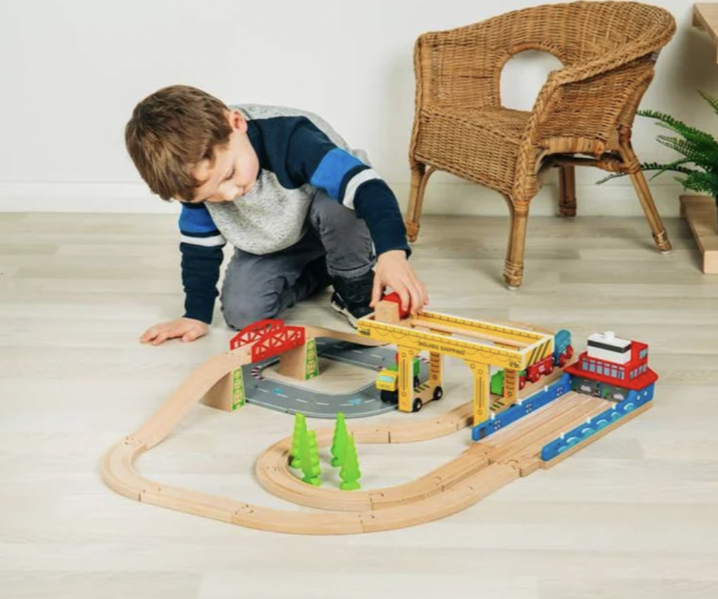 A young boy wearing a blue, gray, and white long-sleeve shirt kneels on a light wood floor to play with a wooden toy train set. The set includes a red train, yellow bridge, and curved tracks.