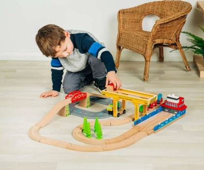 A young boy wearing a blue, gray, and white long-sleeve shirt kneels on a light wood floor to play with a wooden toy train set. The set includes a red train, yellow bridge, and curved tracks.