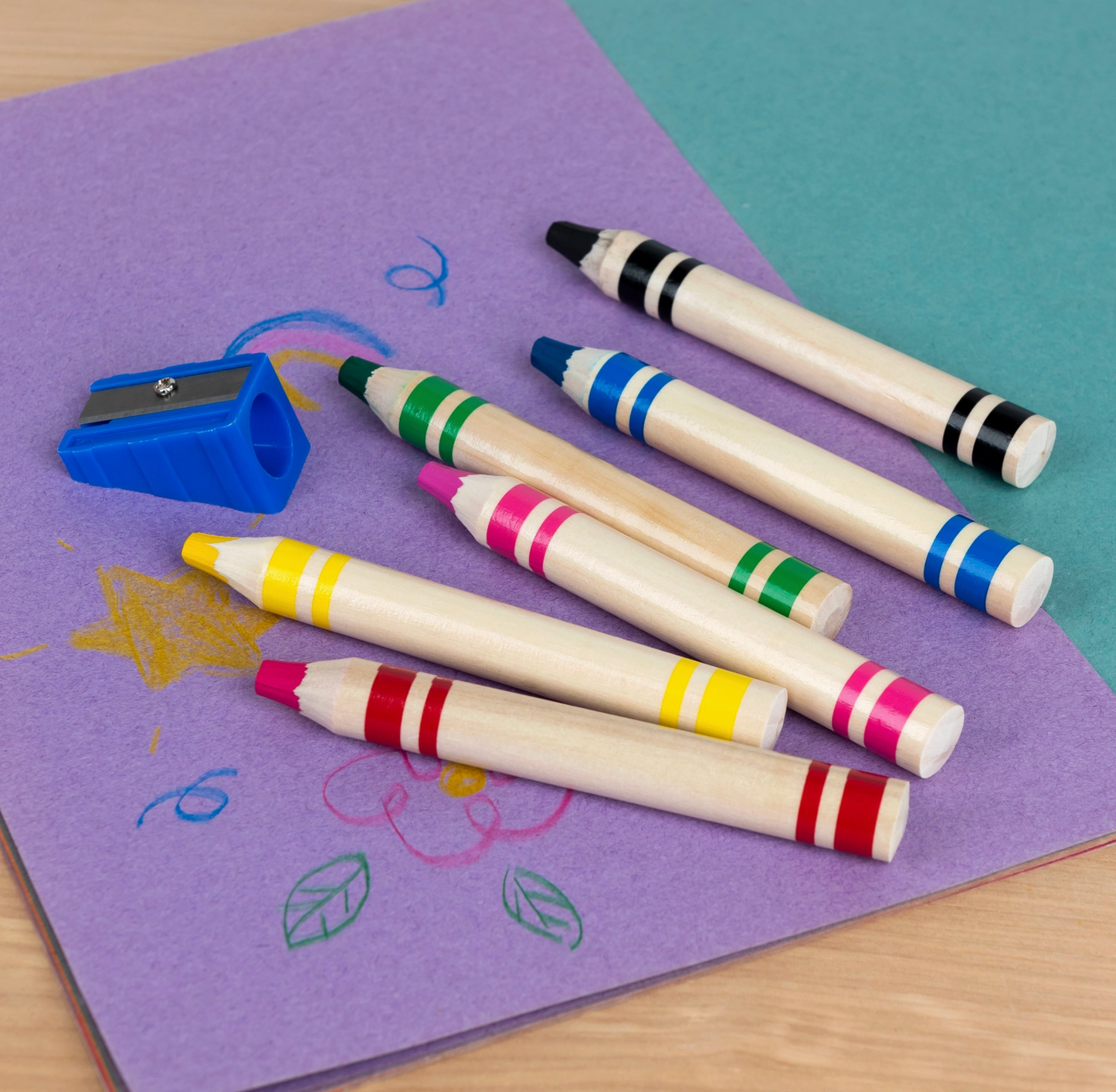 Six Rex London jumbo wooden crayons and blue sharpener laid out on purple paper with children's drawings
