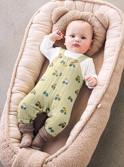 A baby lying on their back inside a beige muslin bassinet, wearing a sage green quilted overalls romper with a tractor pattern, a white long-sleeve shirt, and brown socks.