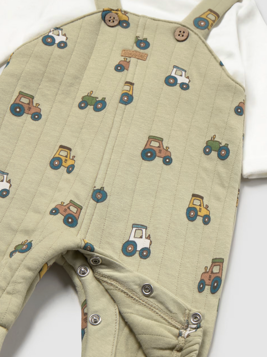 Close-up of a baby's quilted overalls in a light sage green color, featuring an all-over tractor pattern in yellow, green, blue, and orange. The overalls are styled over a white long-sleeve shirt, with snap buttons visible on the chest and legs.