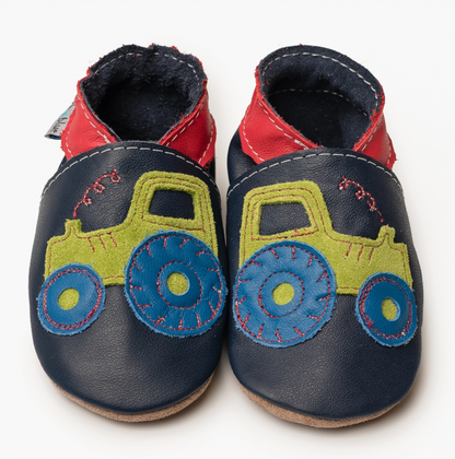 Upper: deep navy blue smooth leather — a strong, rich navy, not a pale blue
Collar/ankle band: bright red leather — bold contrast, very striking; white topstitching runs along the collar seam joining navy to red
Tractor appliqué: the centrepiece — a lime/olive green suede tractor body stitched onto the navy upper; the tractor is a classic farm tractor silhouette with cab, body and three wheels visible; it faces toward the toe
Wheels: bright cobalt/teal blue leather circles — large rear wheel dominating the 