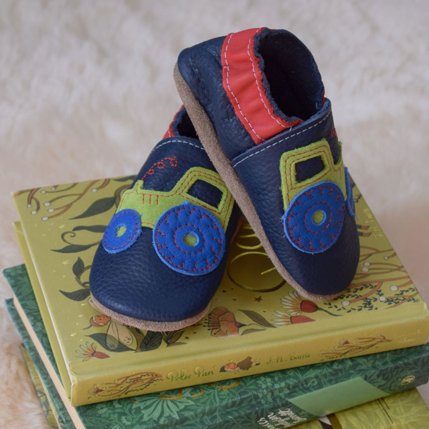Pair of Inch Blue tractor leather baby shoes balanced on top of a small stack of illustrated children's books with yellow-green and green covers featuring botanical and nature illustrations; soft cream textured rug or sheepskin surface in the background; navy blue leather upper clearly visible with lime green suede tractor appliqué and cobalt blue wheels with yellow hub circles and red running stitch tread; bright red leather ankle collar with white topstitching; natural suede sole visible on the bottom sho