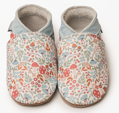 Pair of Inch Blue Winnie handmade leather baby shoes photographed from above against a white background; cream leather upper with an all-over woodland print featuring red and white spotted toadstools, blue cornflower daisy-type flowers with dark centres, small pink and coral five-petal wildflowers, red berry clusters on stems, sage and olive green leaves and orange autumn leaves scattered across the cream ground; contrasting pale powder blue leather ankle collar; fluffy natural suede cream lining visible at