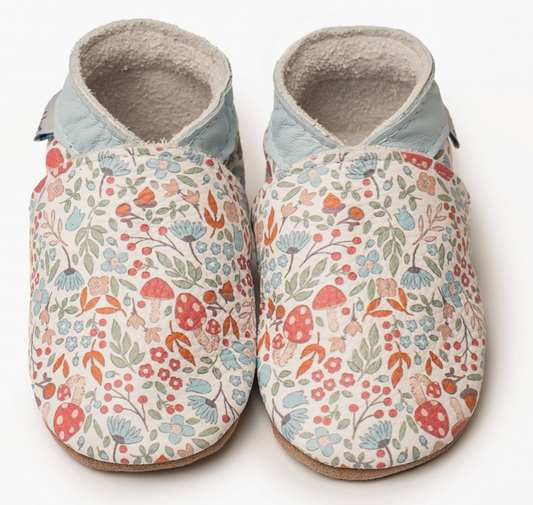 Pair of Inch Blue Winnie handmade leather baby shoes photographed from above against a white background; cream leather upper with an all-over woodland print featuring red and white spotted toadstools, blue cornflower daisy-type flowers with dark centres, small pink and coral five-petal wildflowers, red berry clusters on stems, sage and olive green leaves and orange autumn leaves scattered across the cream ground; contrasting pale powder blue leather ankle collar; fluffy natural suede cream lining visible at