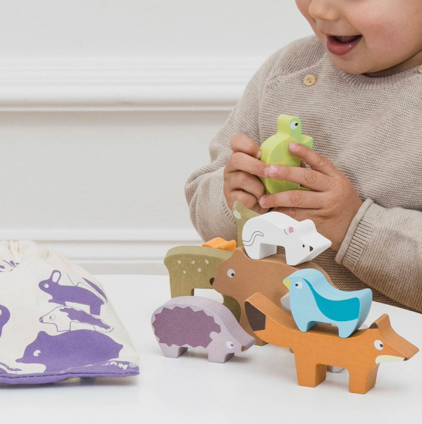 Le Toy Van Forest Stacking Wooden Animals set — lifestyle image of a toddler holding the green tortoise piece and stacking the animals on a white table, with the purple-printed cotton drawstring bag beside them