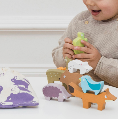 Le Toy Van Forest Stacking Wooden Animals set — lifestyle image of a toddler holding the green tortoise piece and stacking the animals on a white table, with the purple-printed cotton drawstring bag beside them