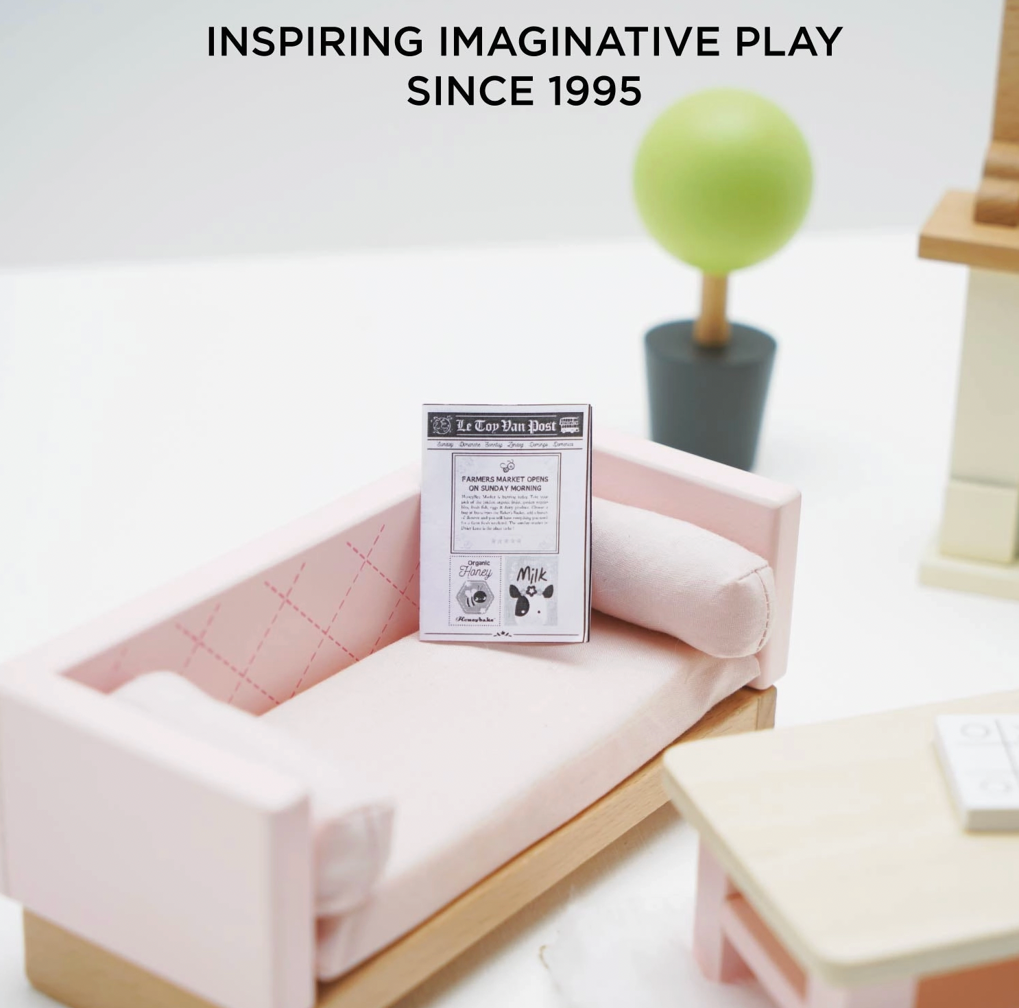 Le Toy Van sitting room dolls house furniture set — close-up lifestyle detail showing the blush pink quilted sofa with bolster cushion and miniature Le Toy Van Post newspaper, with round topiary tree in dark grey pot, "Inspiring Imaginative Play Since 1995" text