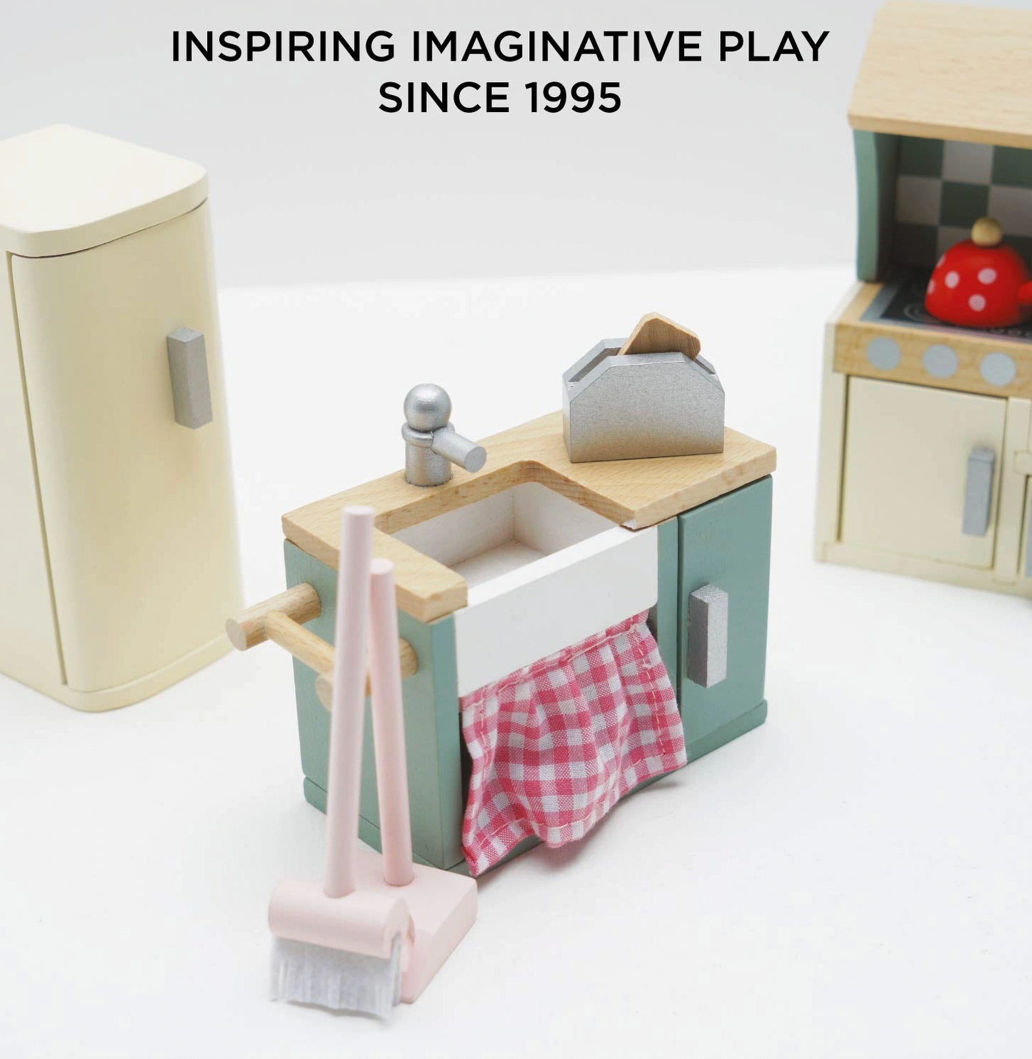 Le Toy Van kitchen dolls house furniture set — close-up detail of the sage green sink unit with white Belfast-style sink, silver tap, pink gingham curtain below and miniature toaster on the worktop; cream retro fridge-freezer and checkerboard splashback cooker visible in background; "Inspiring Imaginative Play Since 1995" heading