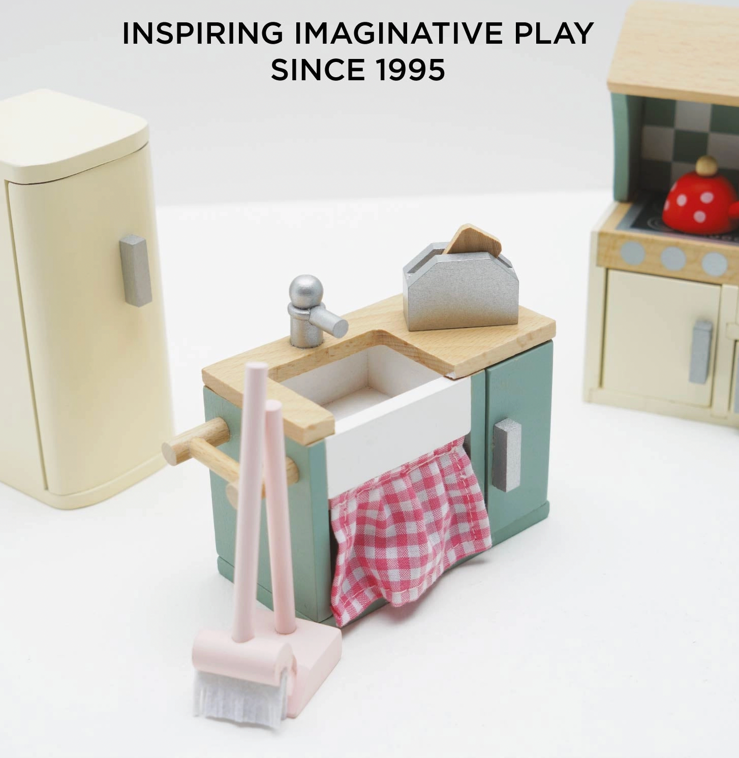 Le Toy Van kitchen dolls house furniture set — close-up detail of the sage green sink unit with white Belfast-style sink, silver tap, pink gingham curtain below and miniature toaster on the worktop; cream retro fridge-freezer and checkerboard splashback cooker visible in background; "Inspiring Imaginative Play Since 1995" heading