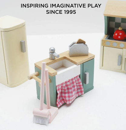 Le Toy Van kitchen dolls house furniture set — close-up detail of the sage green sink unit with white Belfast-style sink, silver tap, pink gingham curtain below and miniature toaster on the worktop; cream retro fridge-freezer and checkerboard splashback cooker visible in background; "Inspiring Imaginative Play Since 1995" heading