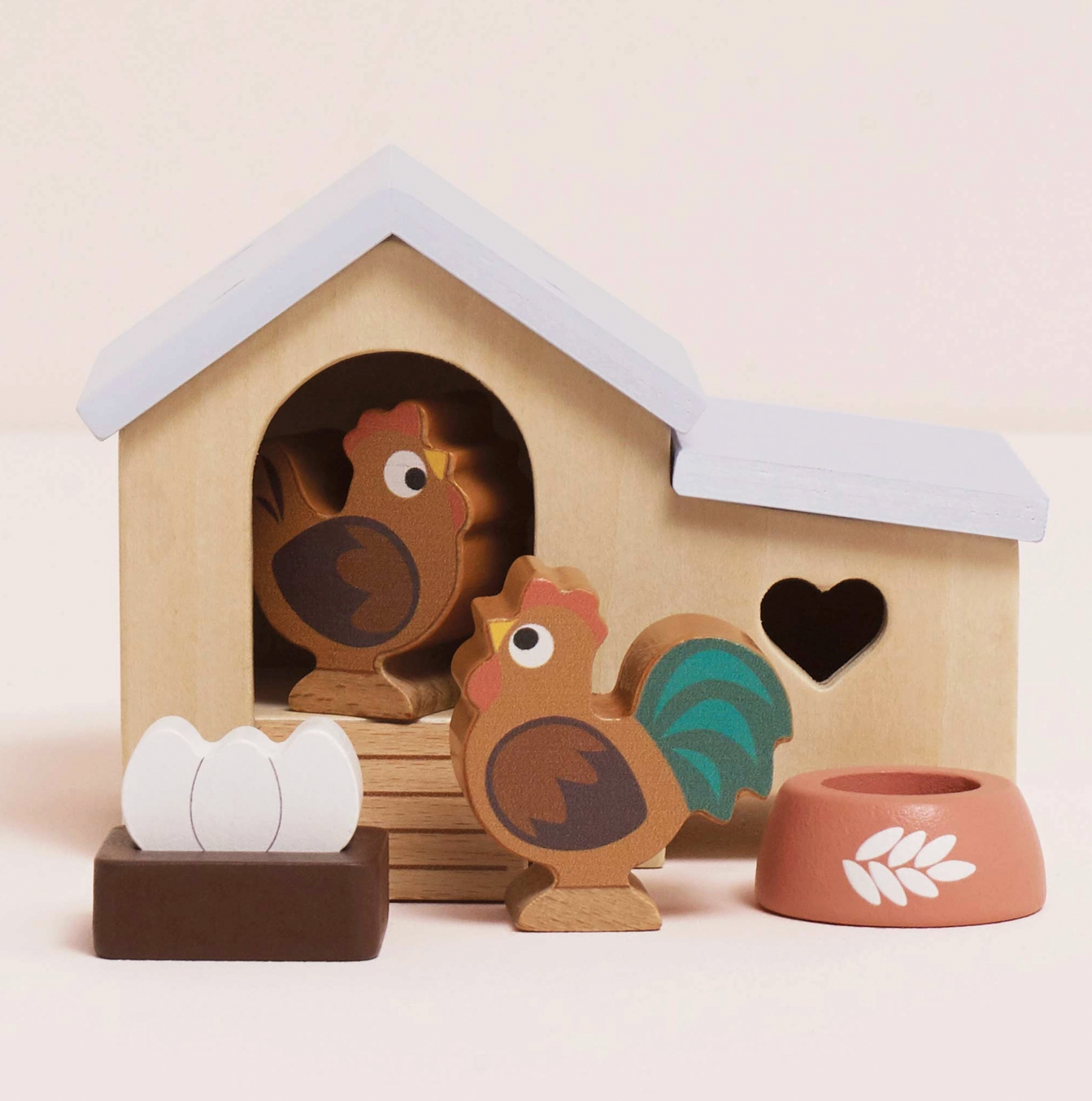 Le Toy Van 6-piece wooden chicken coop set — styled scene showing the natural wood coop with pale blue roof, heart cut-out windows and arched doorway, with rooster figure with teal tail feathers, hen figure, dark brown nest box with two white eggs and terracotta-pink food bowl with white wheat motif