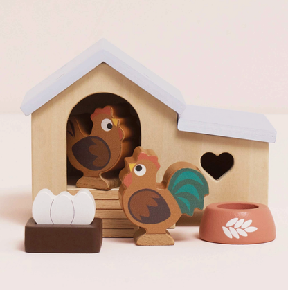 Le Toy Van 6-piece wooden chicken coop set — styled scene showing the natural wood coop with pale blue roof, heart cut-out windows and arched doorway, with rooster figure with teal tail feathers, hen figure, dark brown nest box with two white eggs and terracotta-pink food bowl with white wheat motif