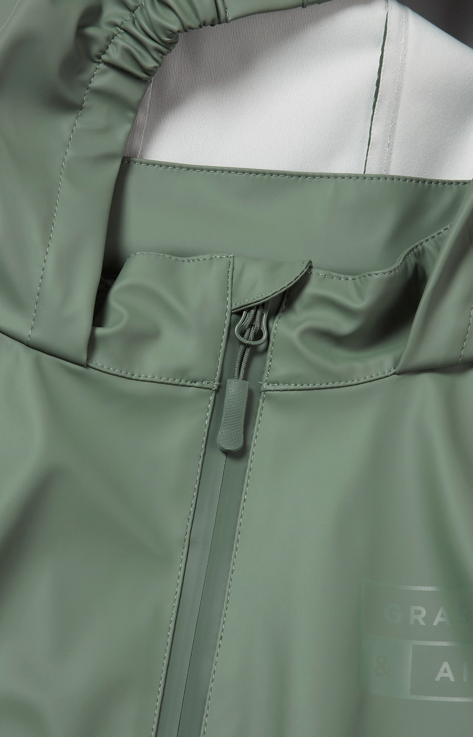 Grass & Air children's waterproof puddle suit — close-up detail of the waterproof front zip with protective zip guard flap and tone-on-tone Grass & Air logo on the chest, with attached elasticated hood visible above