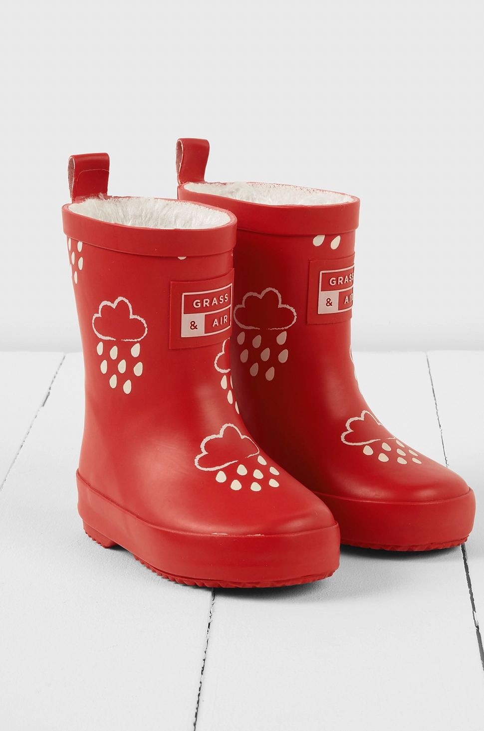 Grass & Air children's ruby red colour-changing wellies — dry state showing rich ruby red natural rubber with cream cloud-and-raindrop motifs, soft cream fleece lining visible at top, two pull-on loops at back and Grass & Air logo badge on side