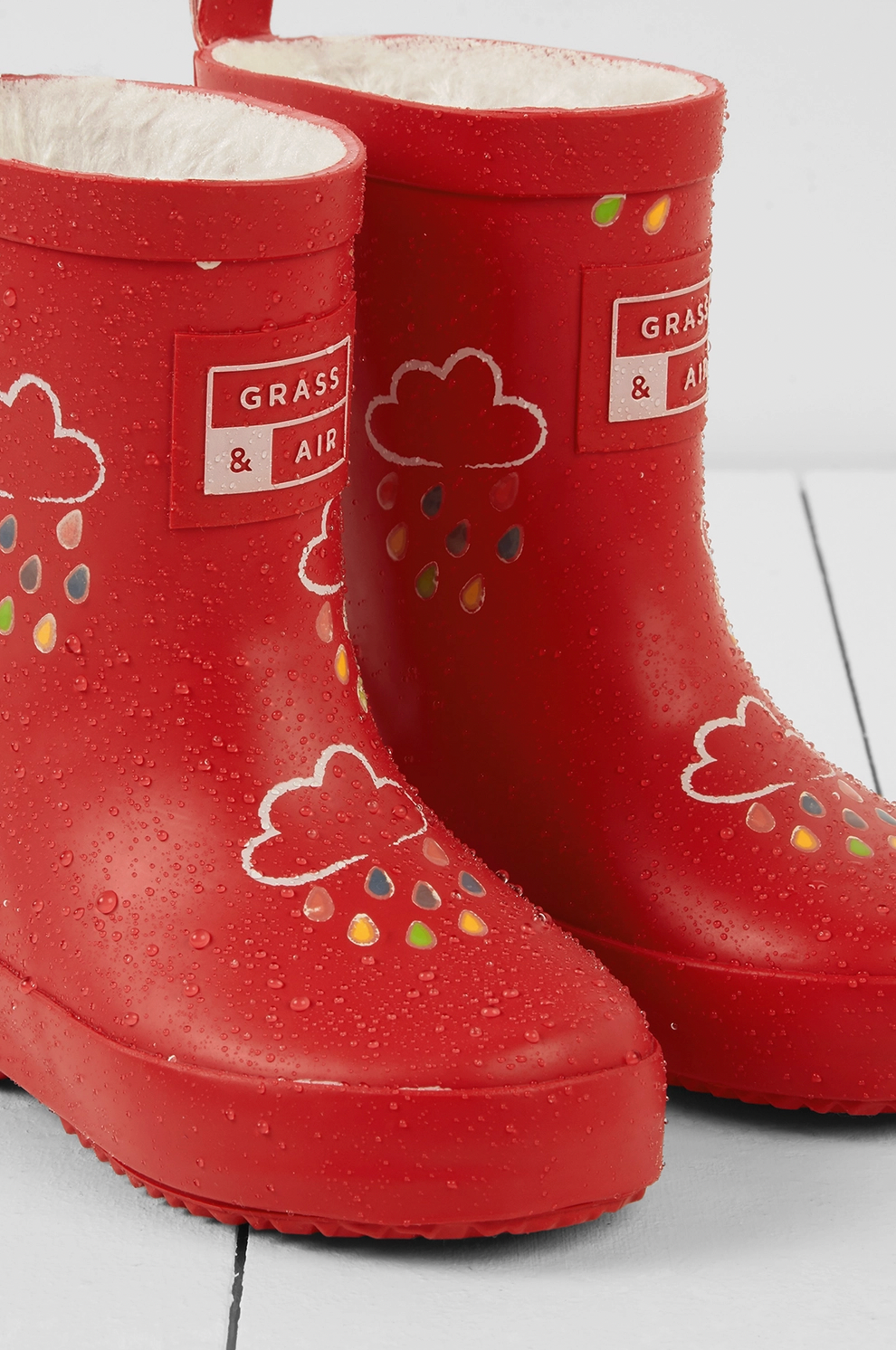 Grass & Air children's ruby red colour-changing wellies — wet state close-up showing the colour-change effect with cream cloud motifs and raindrops revealed in full multicolour (red, orange, yellow, green, blue and teal) as water droplets cover the boots
