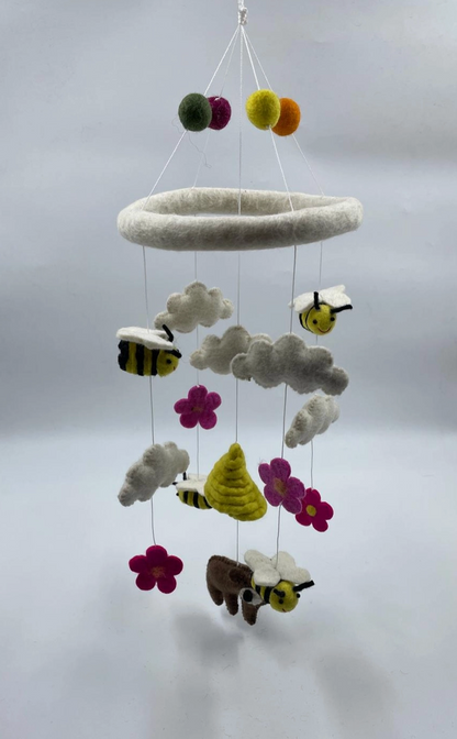 Funky Yak handmade needle-felted bears and bees nursery cot mobile — full view showing a white needle-felted ring hoop with a cluster of multicolour felt pom-pom balls (green, purple, yellow and orange) at the top; suspended on fine white wire at staggered heights are three yellow-and-black striped bees with white wings, one brown bear with white wings, one golden coiled beehive, three soft grey-white felt clouds and three hot pink daisy flowers with yellow centres; approximately 50cm from hoop to lowest po