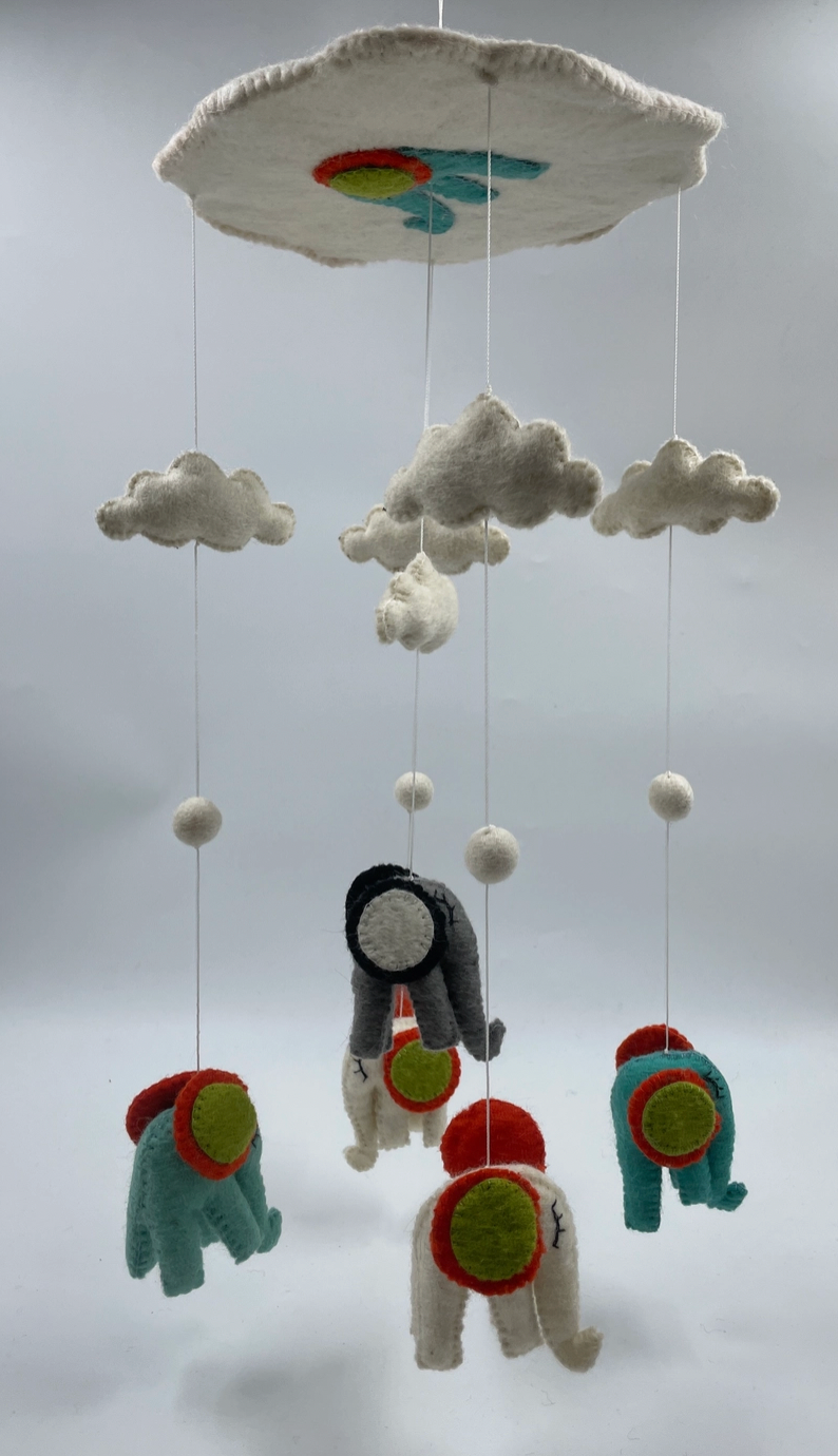 Funky Yak handmade needle-felted elephant nursery cot mobile — low-angle view looking up at the cloud-shaped hoop with teal elephant and yellow-orange sun appliqué, showing the five elephants and white felt clouds suspended below