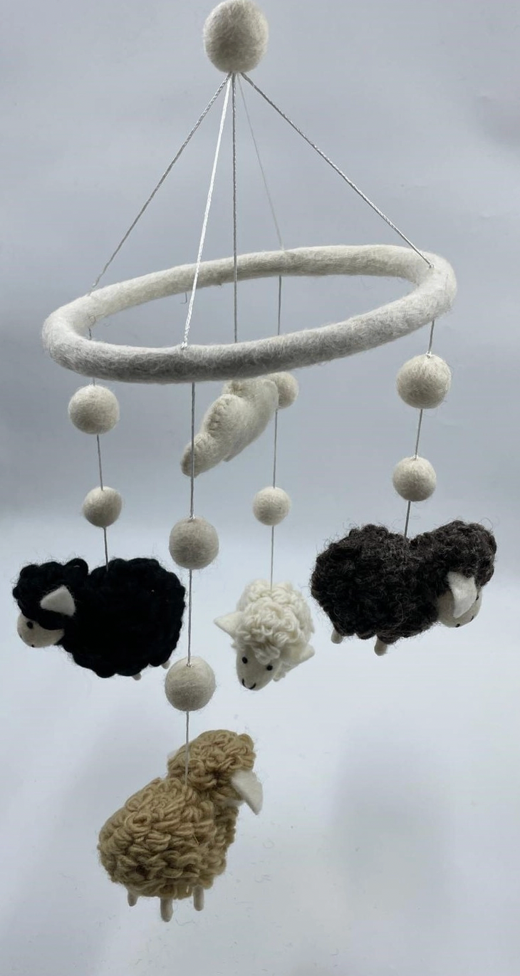 Funky Yak handmade needle-felted sheep nursery cot mobile — five wool felt sheep in four natural fleece colours (jet black, dark charcoal, cream white and sandy caramel) suspended at staggered heights on fine silver wire from a white felt hoop, interspersed with white felt pom-pom balls; single white pom-pom at the top for hanging; approximately 50cm from hoop to lowest point